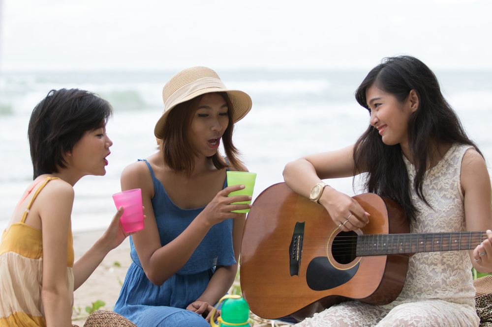 portrait of asian woman and friends group playing guitar and happiness singing a song use for people relaxing holiday at sea side
